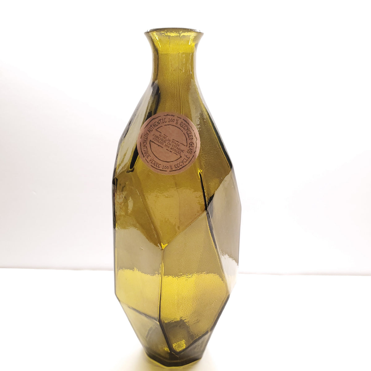 A faceted glass vase with a narrow neck and wide base, made of translucent olive-green glass with a circular paper label attached near the upper middle, photographed against a plain white background.
