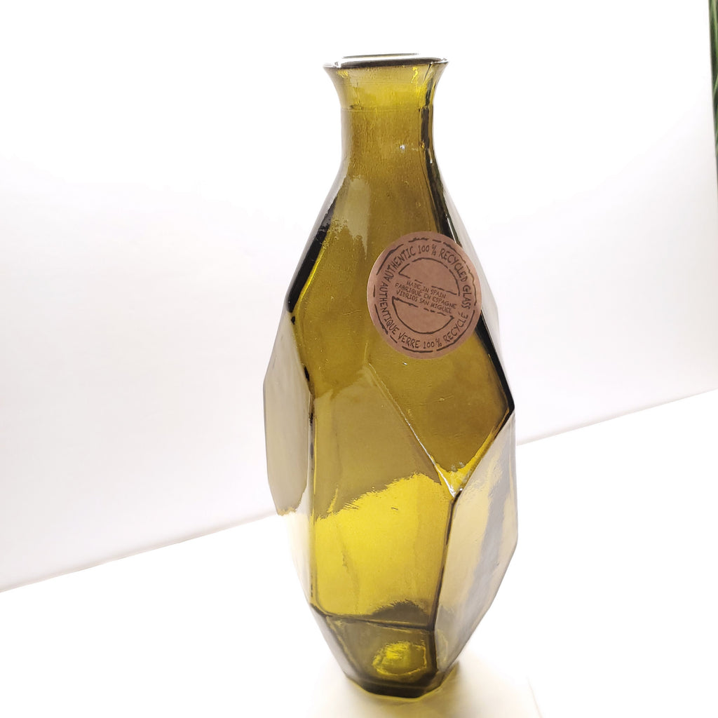 An olive green faceted glass vase with a narrow neck and a circular paper label on the side, photographed against a plain white background.