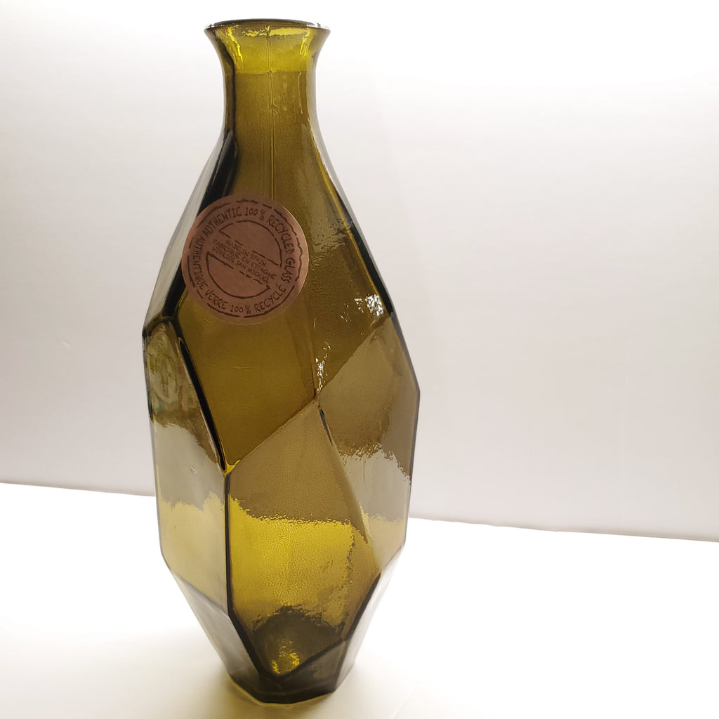 A faceted, geometric glass vase in a translucent olive green color, featuring a circular paper label on the side, set against a plain white background.