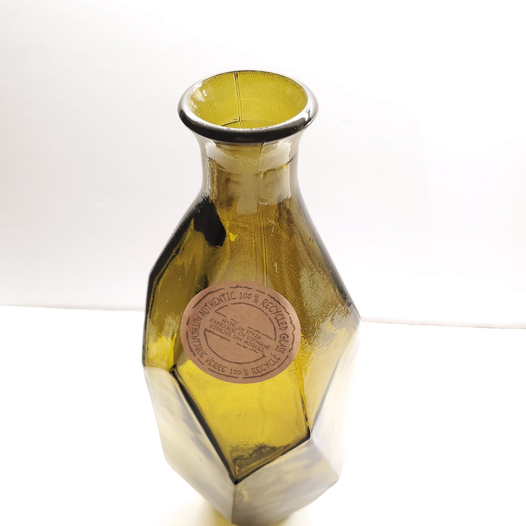 A faceted olive green glass vase with a narrow neck and a circular metallic brand label on the front, photographed against a plain white background.